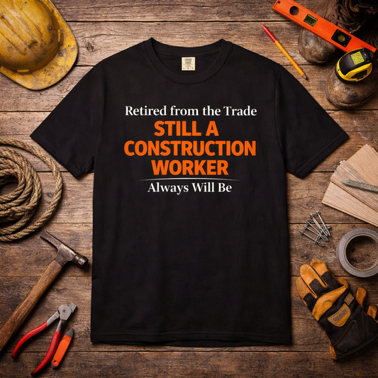Black construction worker retirement T-shirt with orange text 'Retired from the Trade STILL A CONSTRUCTION WORKER Always Will Be' displayed on wooden background with tools