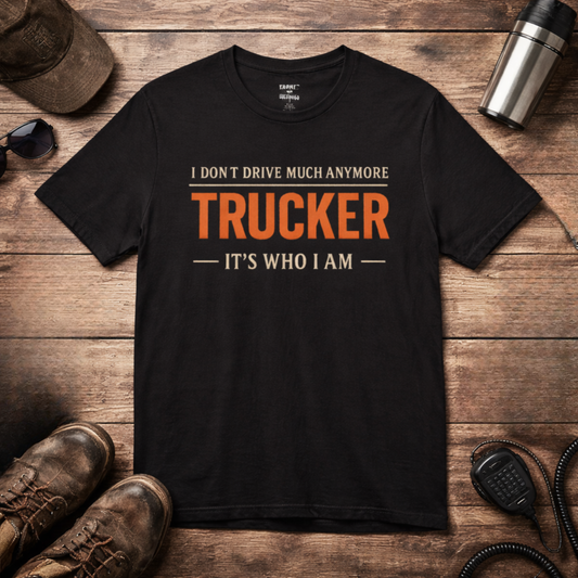 Black retired trucker T-shirt with 'I don't drive much anymore TRUCKER It's Who I Am' design on wooden table background with accessories
